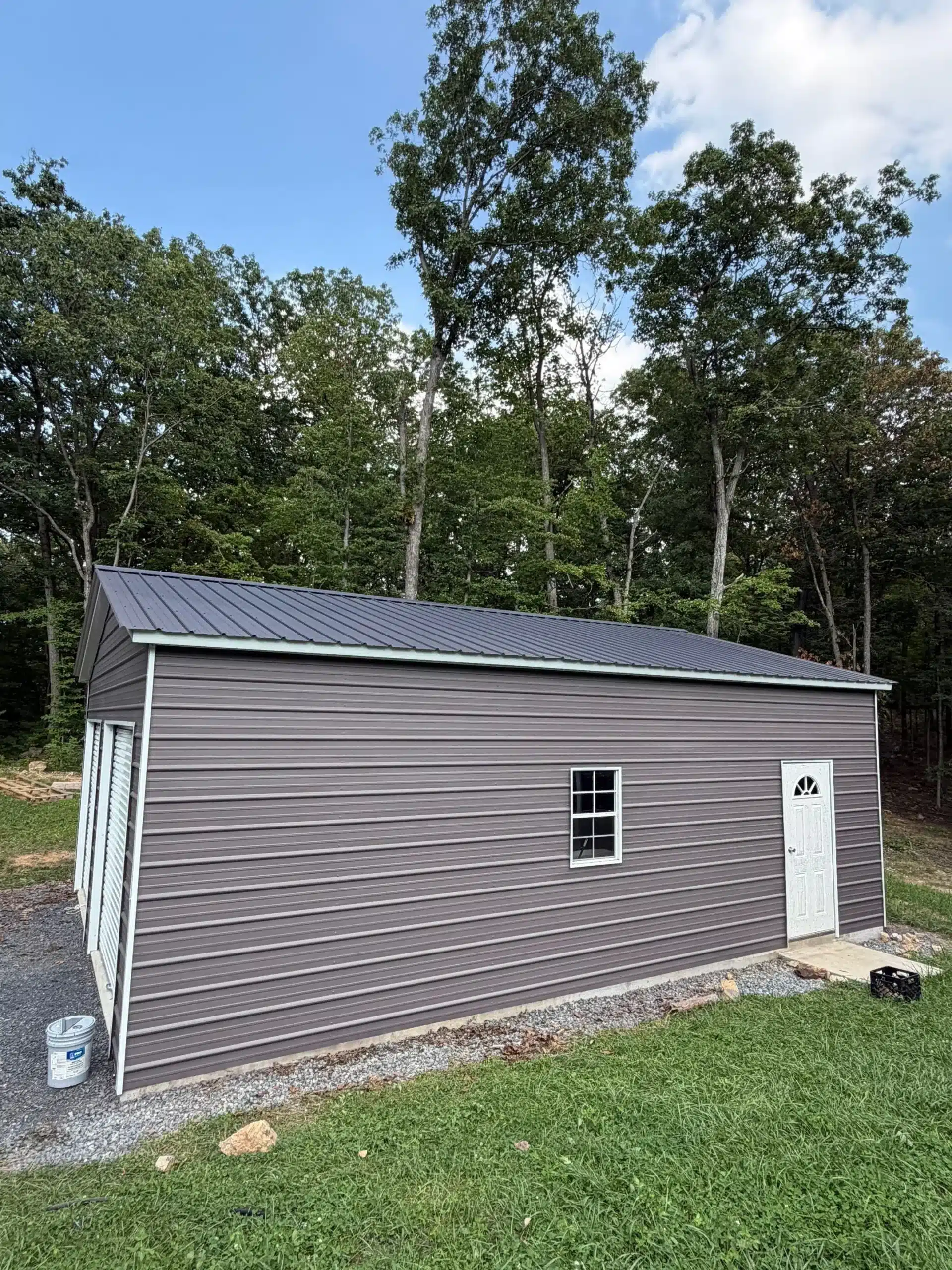 Gray Metal Siding Garage Gray horizontal metal siding garage with white door and window, dark metal roof, gravel pad, and tall trees behind.